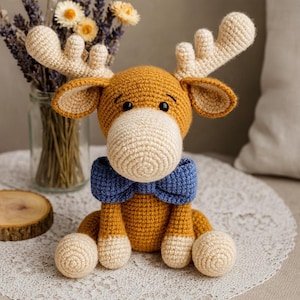 Pode incluir: Um brinquedo de alce de crochê com corpo marrom, chifres e pés creme, e uma gravata borboleta azul. O brinquedo está sobre uma toalha de mesa de renda branca. Um vaso de flores secas está no fundo.