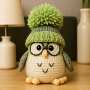 May include: A handmade crochet owl figurine wearing a green knit hat with a pom-pom and glasses. The owl has a cream-colored body, green wings, and yellow feet. The owl is sitting on a wooden surface.