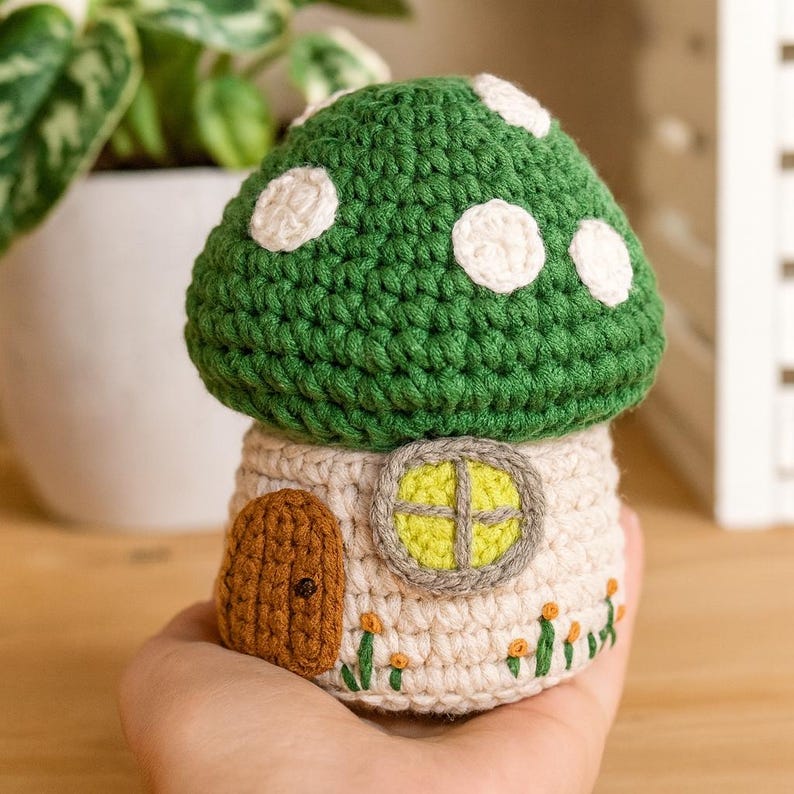 May include: A handmade crochet mushroom house with a green cap and white spots. The house has a tan base with a brown door, a yellow window, and embroidered flowers. A whimsical, decorative item.
