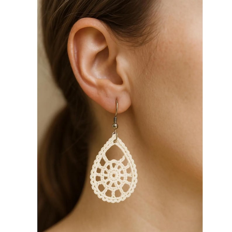 May include: Cream-colored, teardrop-shaped crochet earrings. The earrings feature an openwork design with a central circular pattern and a scalloped edge. The earrings are suspended from a simple metal hook.