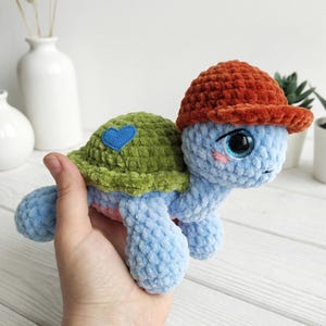 May include: A handmade blue plush turtle with a green shell and a small orange hat. The turtle has a blue heart on its shell and sparkly blue eyes. The turtle is being held in a hand.