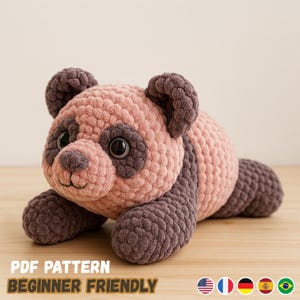 Puede incluir: Un oso panda de ganchillo hecho a mano en tonos rosa y marrón. El peluche tiene grandes ojos negros y está tumbado sobre una superficie de madera. El texto "PDF PATTERN BEGINNER FRIENDLY" se muestra en la parte inferior de la imagen.