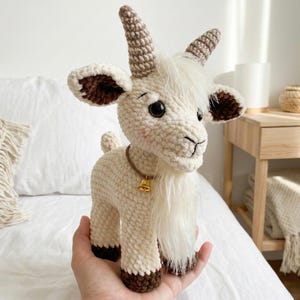 May include: A handmade crochet goat figurine in cream, brown, and white. The goat has large black eyes, a long white beard, and brown horns. It wears a brown collar with a small gold bell. The goat is approximately 18 cm tall.