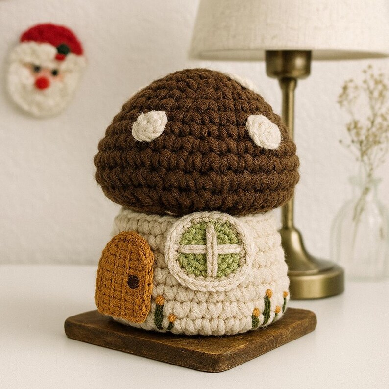May include: A handmade crochet mushroom house with a brown cap and white spots. The house has a cream-colored base with a small orange door and a green and cream window. It sits on a small wooden base. A Santa Claus ornament is in the background.
