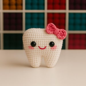 Peut inclure: Une dent crochetée avec un nœud rose, des yeux en bouton noirs et des joues roses. La dent a une bouche souriante et est faite de fil blanc. L'arrière-plan est une grille colorée.
