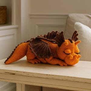Könnte beinhalten: Ein gehäkelter Drache in Orange- und Brauntönen, der mit geschlossenen Augen liegt. Das handgefertigte Plüschtier hat detaillierte Flügel, Hörner und einen stacheligen Rücken. Der Drache liegt auf einer hellen Oberfläche.