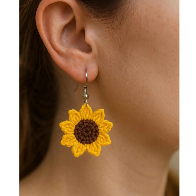 May include: A single yellow and brown crochet sunflower earring is shown. The earring is a detailed replica of a sunflower, with yellow petals and a brown center. The earring is suspended from a silver-colored hook.