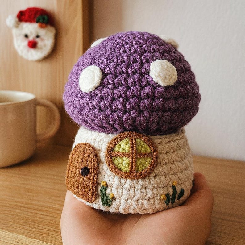 May include: A handmade crochet mushroom house with a purple cap and white spots. The base is cream-colored with a brown door and a round window with green and yellow accents. A Santa Claus face ornament is in the background.