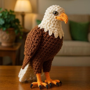 Può includere: Una statuetta di aquila all'uncinetto fatta a mano. L'aquila ha la testa e il collo color crema, il corpo marrone e le piume della coda bianche. Presenta un becco e zampe arancioni con artigli neri. L'aquila è su una superficie di legno.