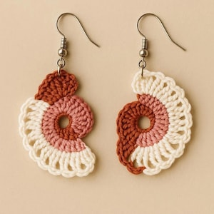 May include: A pair of handmade crochet earrings. Each earring features a spiral design with a color gradient from cream to pink to rust. The earrings have silver-colored fish hook ear wires.