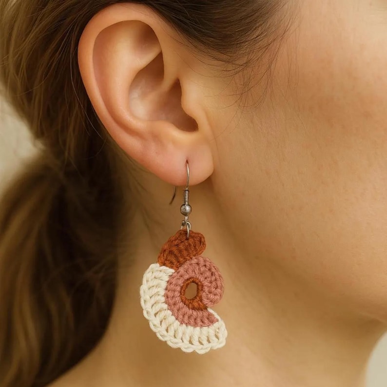 May include: Crocheted spiral earrings in shades of brown, pink, and cream. The earrings are suspended from silver-colored hooks. The design features a spiral shape with a textured, handmade appearance.