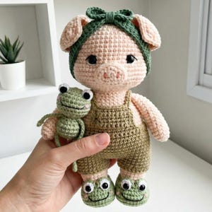 Könnte beinhalten: Gehäkelte Schweinepuppe mit grünen Latzhosen, einem grünen Stirnband und Schuhen mit Froschmotiv. Das Schwein hält einen kleinen grünen gehäkelten Frosch. Die Puppe ist aus hellrosa und grüner Wolle gefertigt.
