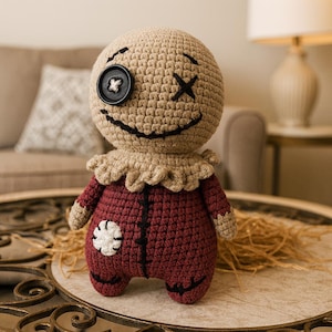 May include: Crocheted doll with a tan head, black stitched eyes, and a button eye. The doll has a burgundy body with a white flower patch and tan arms. It has a ruffled collar and a stitched smile. The doll is sitting on a decorative table.