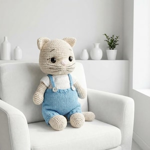 May include: A crocheted cat doll with a cream-colored head and body, wearing a white shirt and light blue overalls. The cat has large black eyes, a pink nose, and black whiskers. It is sitting on a white armchair.