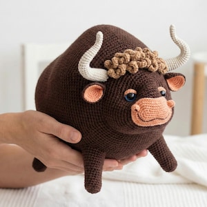 May include: A handmade, crocheted bull plush toy in shades of brown, tan, and cream. The bull has curved cream-colored horns, blue eyes, and a tan muzzle and ears. The plush is held in a person's hands against a soft, white background.