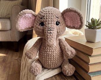 Puede incluir: Un peluche de elefante tejido a mano en tonos marrón y rosa. El elefante tiene orejas grandes con el interior rosa, una larga trompa y ojos negros pequeños. Está sentado sobre una superficie de madera, con una manta suave debajo.