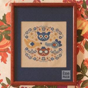 May include: A framed cross-stitch embroidery featuring a circular design with four animal faces: a blue owl, a brown bear wearing a hat, and two birds. The design is surrounded by a floral border and the text "Éire Póca" is in the lower right corner.