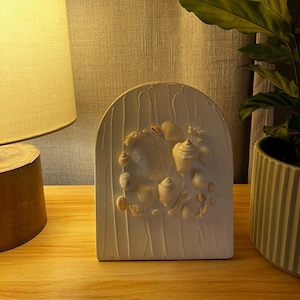 May include: White ceramic bookend shaped like an arch with a raised design of seashells. The bookend is on a wooden surface with a lamp and a potted plant in the background.