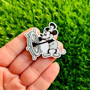 May include: A cartoon sticker featuring Mickey Mouse in a sailor's outfit, holding a ship's wheel. The sticker has a white border and is set against a green background. The character is in black, white, and light blue.