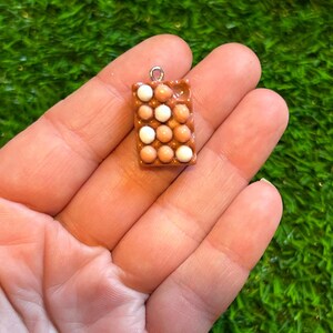 May include: A small, rectangular charm with a pattern of brown and white spheres, similar to a carton of eggs. The charm has a silver loop at the top for hanging. The charm is held in a person's hand.