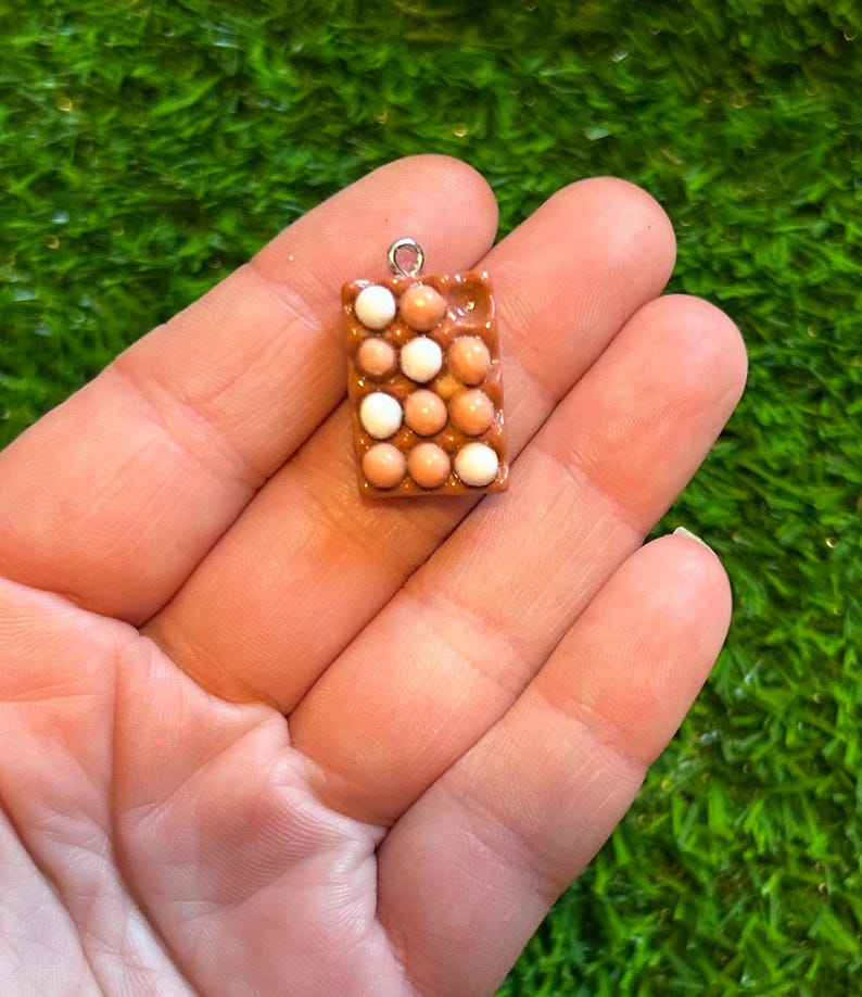 May include: A small, rectangular charm with a pattern of brown and white spheres, similar to a carton of eggs. The charm has a silver loop at the top for hanging. The charm is held in a person's hand.