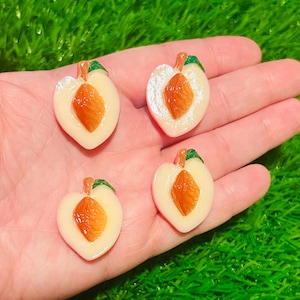 May include: Four miniature peach slice charms. Each charm features a cream-colored peach flesh with a brown pit, a green leaf, and an orange stem. The charms are held in a hand against a green background.