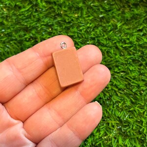 May include: A small, rectangular, light brown charm with a silver-coloured loop at the top. The charm is held in a person's hand against a backdrop of green artificial grass. The charm appears to be made of a solid material.