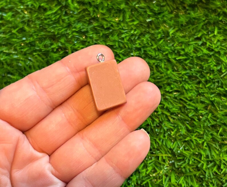 May include: A small, rectangular, light brown charm with a silver-coloured loop at the top. The charm is held in a person's hand against a backdrop of green artificial grass. The charm appears to be made of a solid material.