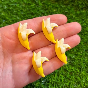 Op de afbeelding: Vier gele bananenbedels met witte binnenkant. De bananen zijn geschild en tonen het witte fruit. De bedels zijn geplaatst tegen een groene achtergrond.