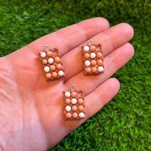 Puede incluir: Tres colgantes rectangulares en miniatura que se asemejan a cartones de huevos. Cada colgante es de color marrón claro con pequeñas cuentas redondas blancas y marrón claro dispuestas en un patrón de cuadrícula. Un lazo plateado está unido a la parte superior de cada colgante.