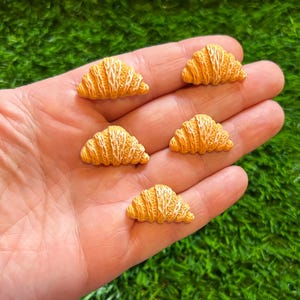 May include: Five miniature, detailed croissants with a golden-brown color and white accents. The miniature food items are arranged on a person's open palm, with a green, grassy background.