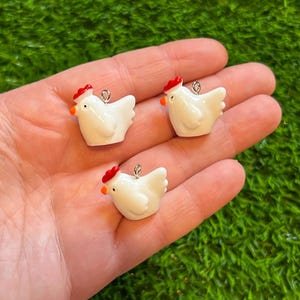 Puede incluir: Tres pequeños colgantes de pollo blancos, con cresta roja y pico naranja. Cada colgante tiene un lazo plateado en la parte superior para colgar. Los colgantes se muestran en una mano sobre un fondo verde.