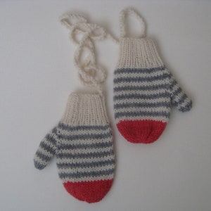May include: A pair of knitted mittens with a cream-colored cuff, gray and white horizontal stripes, and a red bottom. The mittens are connected by a cream-colored string. The mittens are designed for warmth and protection from the cold.