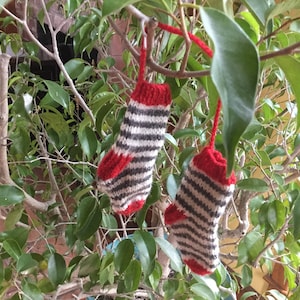 May include: Two miniature knitted Christmas stockings, each with a red cuff, heel, and toe, and a body striped with alternating gray and off-white bands. The stockings are suspended from a tree branch by red yarn loops, set against a backdrop of green leaves.