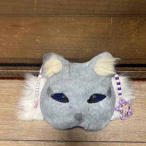 May include: Grey cat mask with white fur trim and purple and pink beaded accents. The mask has blue mesh eyes and the text "COTTON" is written on the left side.