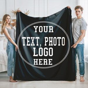 May include: Black fleece blanket with a white circle containing the text "YOUR TEXT, PHOTO LOGO HERE". Two people are holding the blanket.