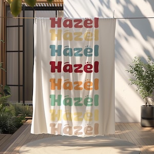 May include: A white beach towel with the name "Hazel" repeated in a rainbow of colors, including red, orange, yellow, green, blue, and purple.