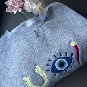 May include: A gray sweatshirt with embroidered designs. The design includes a blue eye with a white and black center, a yellow horseshoe, and a red chili pepper. The sweatshirt is made of a soft material.
