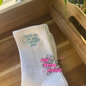 May include: White crew socks with the embroidered text "hold on let me ChatGPT this" in light blue. The socks also feature a pink logo that says "Sew Bossy Threads".