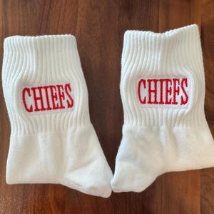 May include: Pair of white ankle socks with the word "CHIEFS" embroidered in red. The socks are made of a soft, ribbed knit material. The socks are likely for sports fans.