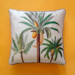 Peut inclure: Un coussin blanc avec une bordure beige présente une illustration colorée de palmiers aux feuilles vertes et aux fruits jaunes.