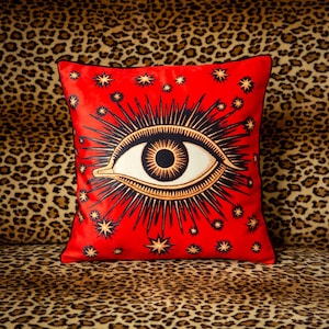 Può includere: Cuscino in velluto rosso con un grande disegno di occhio nero e dorato circondato da stelle dorate. Il cuscino è su uno sfondo leopardato.