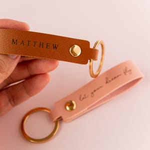 Może przedstawiać: Dwa skórzane breloki z złotymi kółkami. Jeden brelok jest brązowy i ma napis "MATTHEW". Drugi brelok jest różowy i ma napis "Niech twoje marzenie wzbije się w lot".