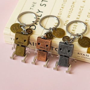 May include: Three robot-shaped keychains with different colored finishes: silver, copper, and brass. Each keychain has a small metal charm with a letter and a phrase. The charms read "live laugh love", "one tiny dream", and "love and beloved".
