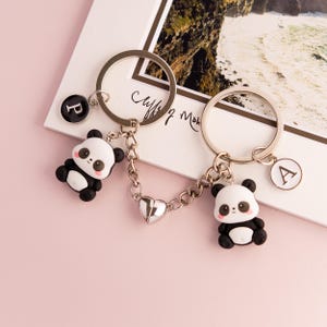Könnte beinhalten: Ein Set aus zwei silbernen Schlüsselanhängern mit Pandabär-Anhängern. Die Schlüsselanhänger sind durch eine silberne Kette mit einem Herz-Anhänger verbunden. Die Schlüsselanhänger haben Buchstaben-Anhänger mit den Buchstaben "P" und "A".