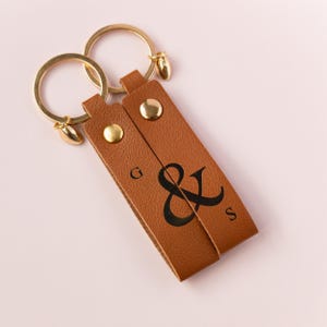 Może przedstawiać: Brązowy skórzany brelok do kluczy z dwoma złotymi kółkami na klucze i złotym zawieszką w kształcie serca. Brelok ma wygrawerowane litery "G & S" i symbol ampersanda.