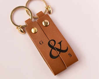 May include: A brown leather keychain with two gold key rings and a gold heart charm. The keychain has the letters "G & S" and an ampersand symbol engraved on it.