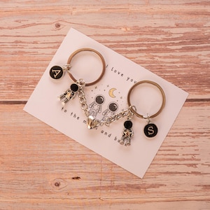 May include: Two silver-toned metal keychains connected by a magnetic heart charm. Each keychain features a small astronaut figure and a circular black charm with the letters A and S in gold. The keychains are displayed on a white card with the text I love you to the moon and back.