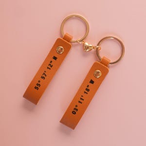 May include: Two orange leather keychains connected by a gold heart-shaped charm. Each keychain features a gold metal ring and a gold rivet. The left keychain is stamped with 55° 57' 12" N and the right keychain is stamped with 03° 11' 18" W.