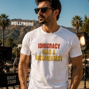 Peut inclure: Un t-shirt blanc à manches courtes avec le texte rouge et jaune "IDIOCRACY WAS A DOCUMENTARY" imprimé sur la poitrine. Le vêtement présente une encolure ronde et une coupe standard, porté avec un pantalon sombre et une montre au poignet.
